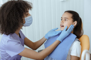 Young serious african american doctor in uniform, protective mask and gloves checks patients mouth